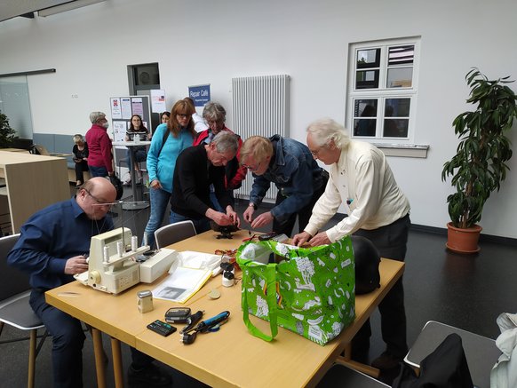 Menschen stehen um einen Tisch und reparieren verschiedene Dinge Menschen stehen um einen Tisch und reparieren verschiedene Dinge